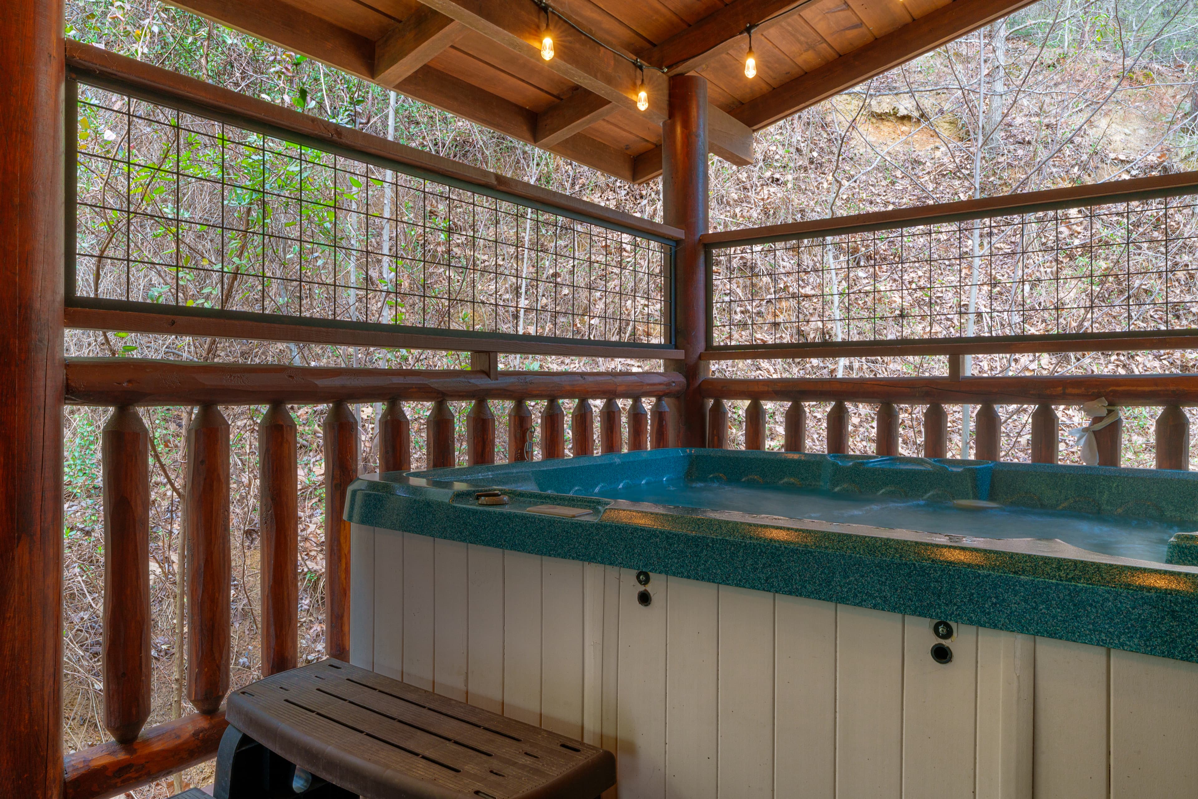 Outdoor hot tub on a rustic wooden porch with string lights and a forest view.
