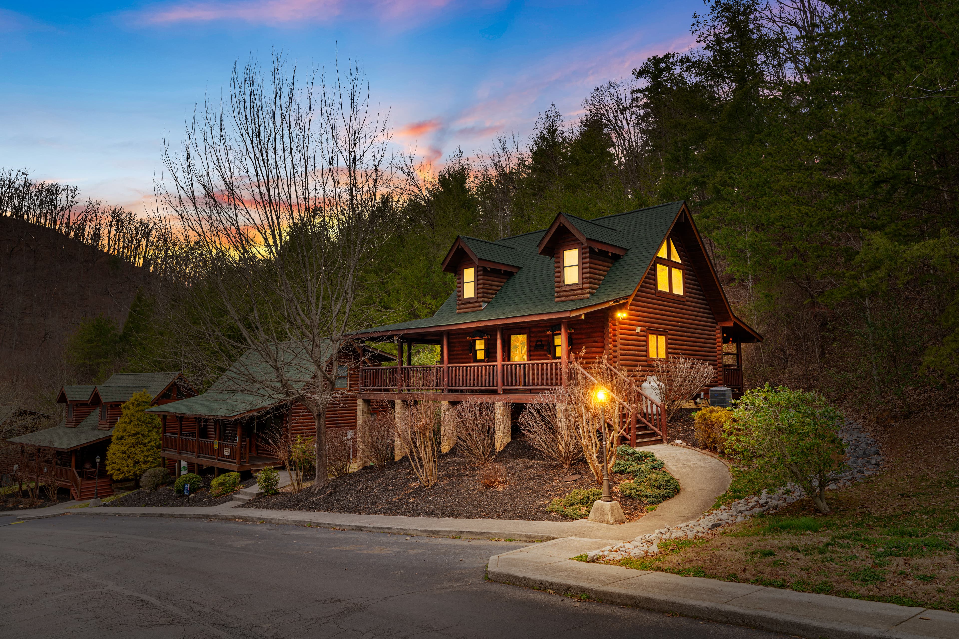 Cozy log cabin with glowing windows on a wooded hillside under a colorful sunset.